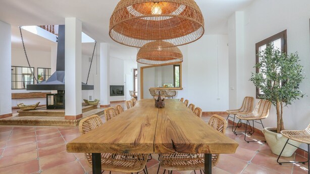 Un vaste espace dédié à la salle à manger, avec une longue table en bois entourée de chaises en osier, un grand luminaire tressé suspendu au-dessus et une plante en pot placée sur la droite.