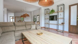 Un salon avec un canapé d’angle beige, une table basse en bois et un téléviseur posé sur un meuble noir. On y trouve des étagères avec des plantes et des objets décoratifs, ainsi qu’un luminaire suspendu en osier.