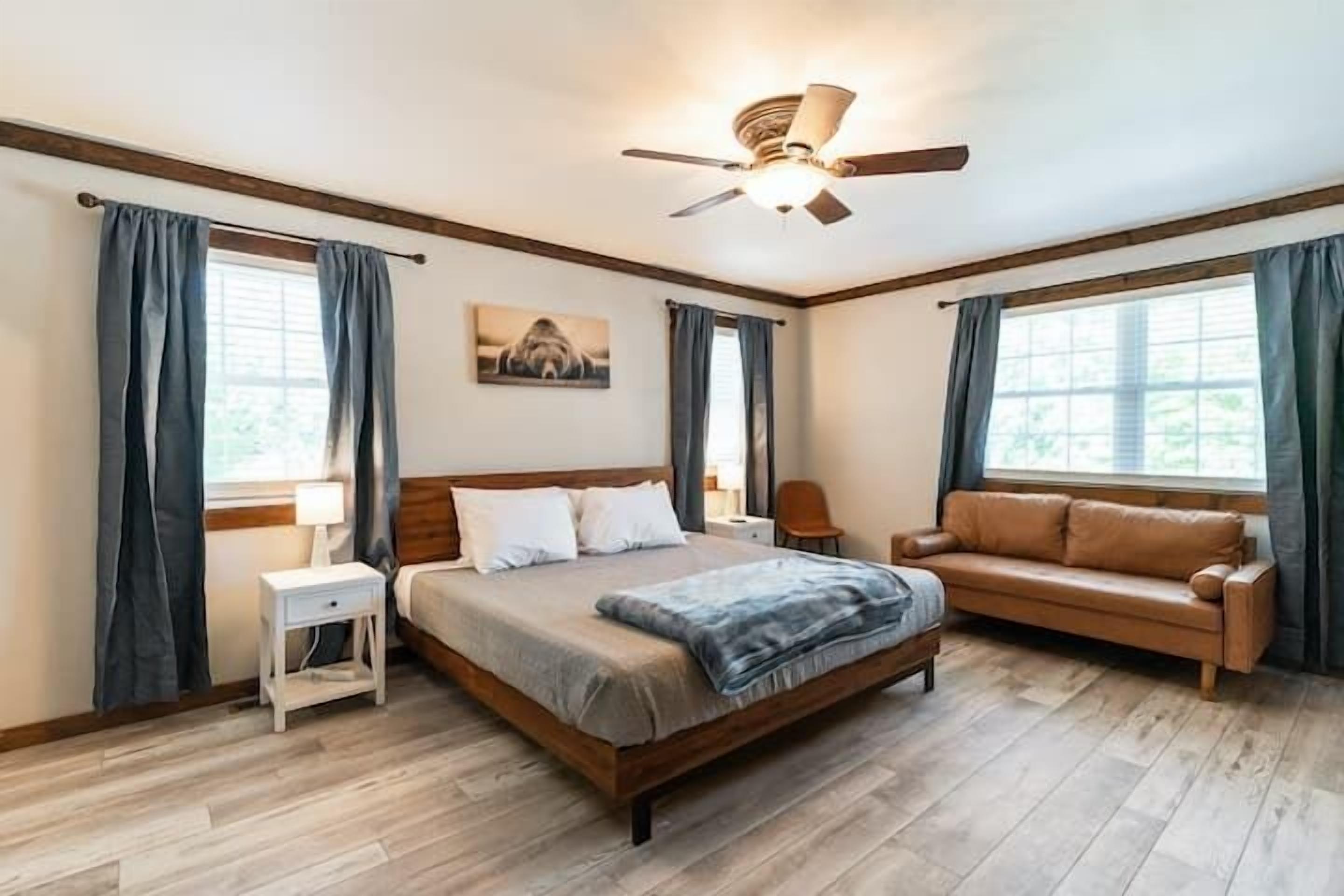 Ein Schlafzimmer mit einem Bett, einem Schlafsofa, einem Nachttisch, einem Deckenventilator und großen Fenstern mit Vorhängen.