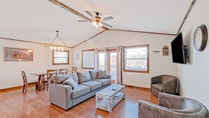 Ein geräumiges Wohnzimmer mit grauem Sofa, einem Couchtisch und einem Deckenventilator. Es gibt einen Essbereich mit Tisch und Stühlen, und eine Uhr an der Wand.