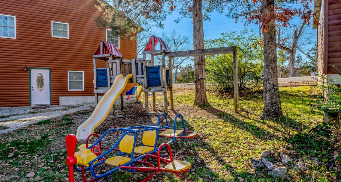 A playground with a slide and swings, surrounded by trees and a wooden building.