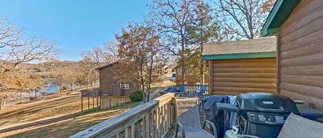A wooden deck with a barbecue grill and chairs, overlooking a hilly landscape with a small building in the background.