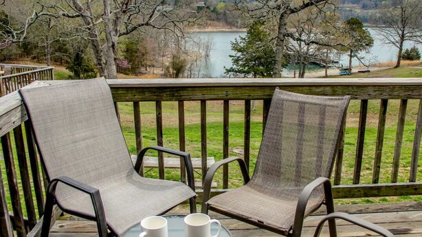 A wooden deck with two chairs and a table, overlooking a lake and trees. - 1BR Waterfront Cabin w/ Pool + Swim & Boat dock (Branson)