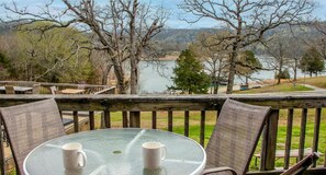 A glass table with two white mugs, surrounded by wicker chairs, overlooking a lake with trees and a wooden deck.