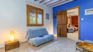 A room with a blue wall, a wooden door, a sofa with a patterned cover, a small table with a lamp, and a tiled floor. - Cortijo Relatillos Albox by Ruralidays (Almería)
