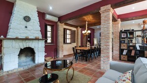 A cozy living room with a stone fireplace, a dining area with a wooden table and chairs, and a bookshelf filled with books and decorative items. - Valdemarín Rural Cortijos Nuevos by Ruralidays (Jaén)