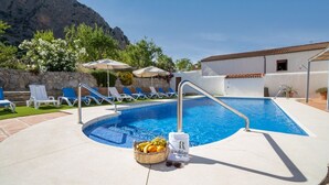 Une piscine avec des chaises longues et des parasols, un panier de fruits et une serviette ornée d’un logo.