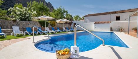 Una piscina con sillas de descanso y sombrillas, una cesta de fruta y una toalla con un logotipo.