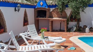 A poolside area with lounge chairs, a table with a vase of flowers, and a brick oven.
