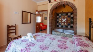 A bedroom with a bed, a chair, a red sofa, a brick archway, and a tiled wall.