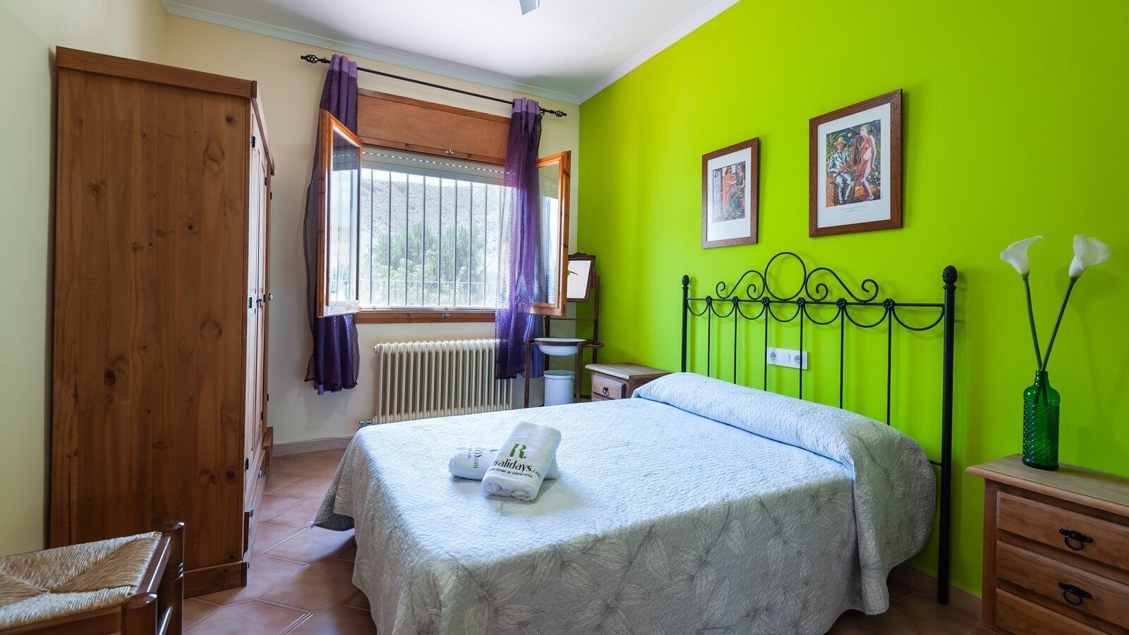 Un dormitorio con cama, armario de madera, ventana con cortinas, radiador y pared verde con cuadros enmarcados.