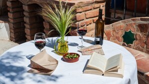 Una mesa redonda con mantel blanco, dos copas de vino, una botella de vino, un florero con follaje verde, un cuenco de aceitunas y un libro abierto.