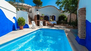 Una piscina con sillones de descanso y un patio, rodeada por un edificio con paredes azules y blancas y un techo de tejas.