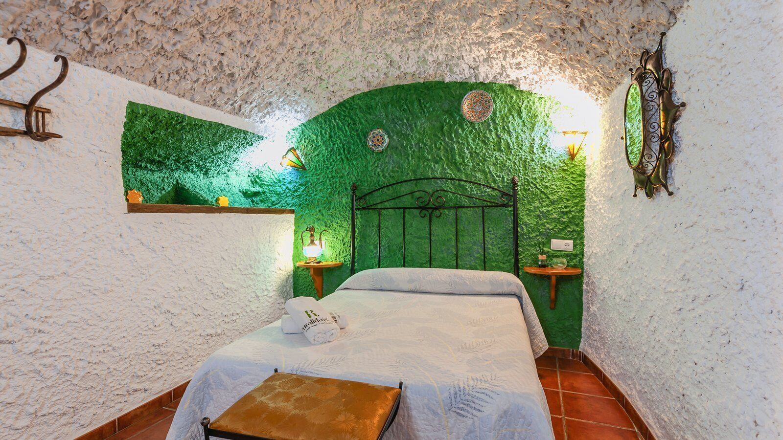A bedroom with a green textured wall, a bed with white linens, a wooden bench, and two bedside tables with lamps.