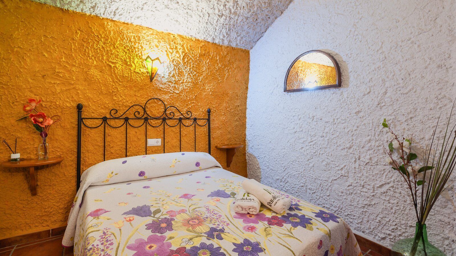 Une chambre à coucher avec un lit, une table de chevet, une lampe fixée au mur et un vase rempli de fleurs.