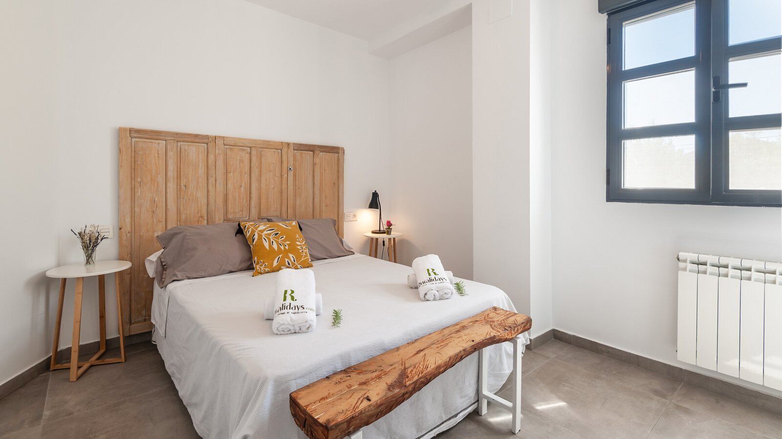 A bedroom with a wooden headboard, a bedside table with a vase, a bench, and two towels with a logo.
