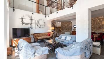 A spacious living area with a high ceiling, a large flat-screen TV, a sofa, a coffee table, and a stone wall in the background.