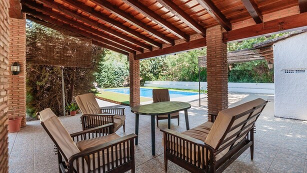A covered patio area with wooden furniture and a pool in the background.