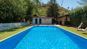 A rectangular swimming pool with clear blue water, surrounded by a white fence and a grassy area. There is a small building with a tiled roof and a door in the background.