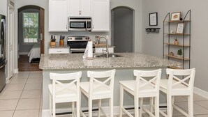 Une cuisine moderne avec des meubles blancs, un plan de travail en granit et un four encastré. Il y a trois chaises blanches autour de l’îlot central.
