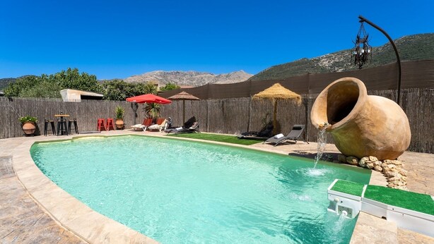 Piscina com um elemento aquático, cercada por espreguiçadeiras e guarda-sóis, com montanhas ao fundo.