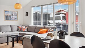 Una sala moderna con un sofá gris, una mesa de centro negra y cojines naranjas. En primer plano, una mesa de comedor con sillas. Amplias ventanas ofrecen vistas de un edificio rojo y vehículos estacionados afuera.