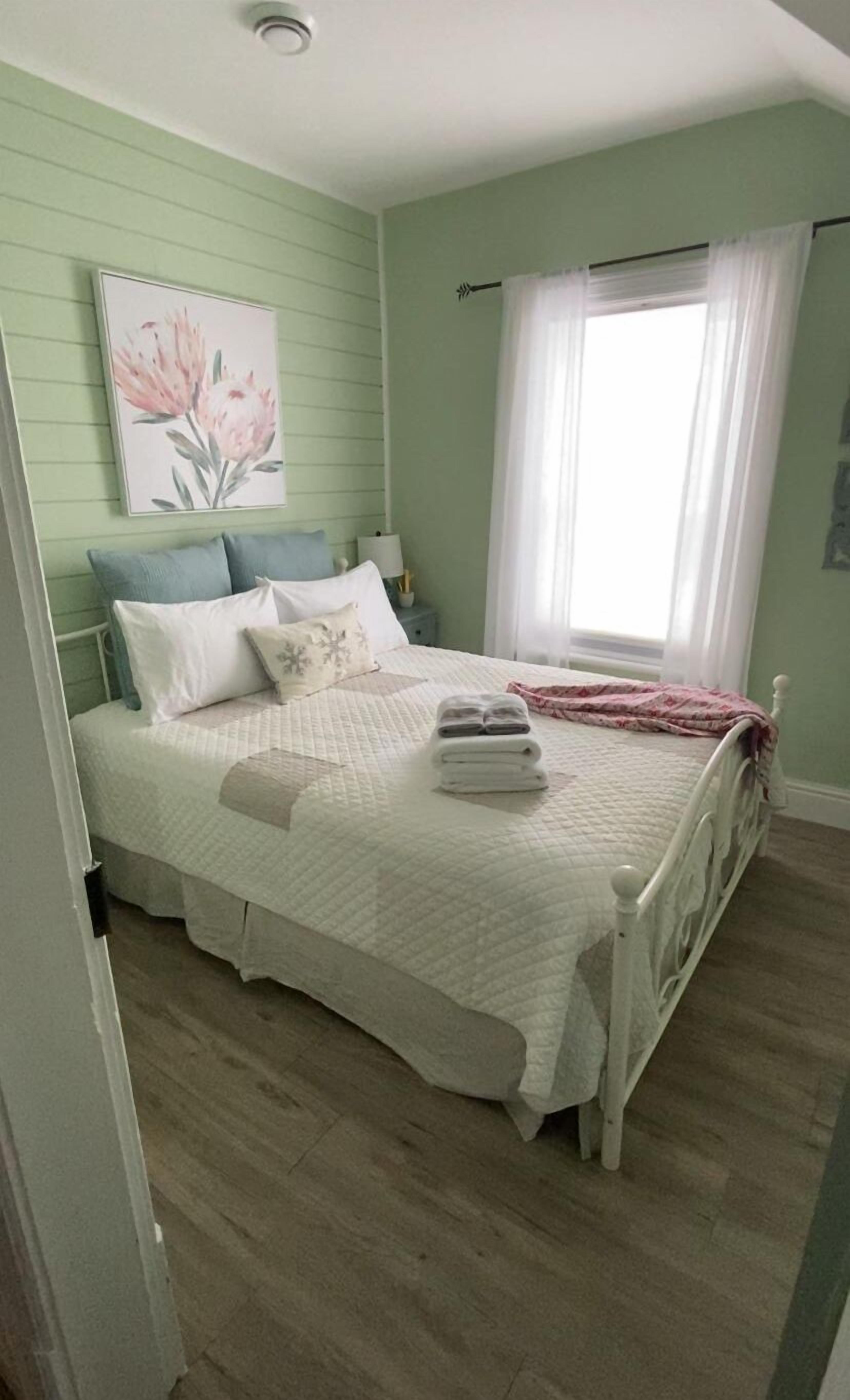 Un dormitorio con cama, almohadas, una mesita de noche y un cuadro floral en la pared.