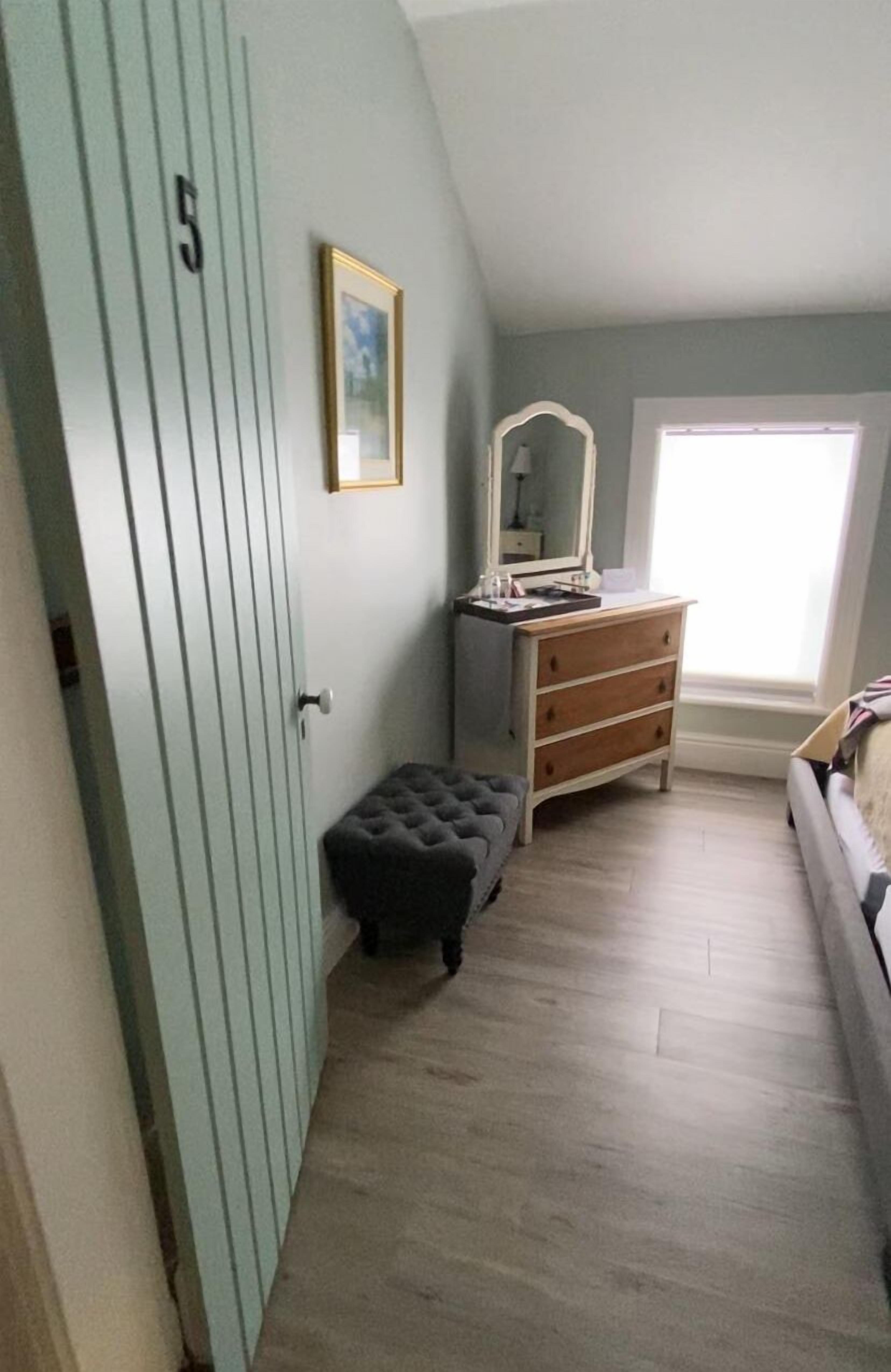 A bedroom with a light blue door, a wooden dresser, a mirror, and a window with natural light.