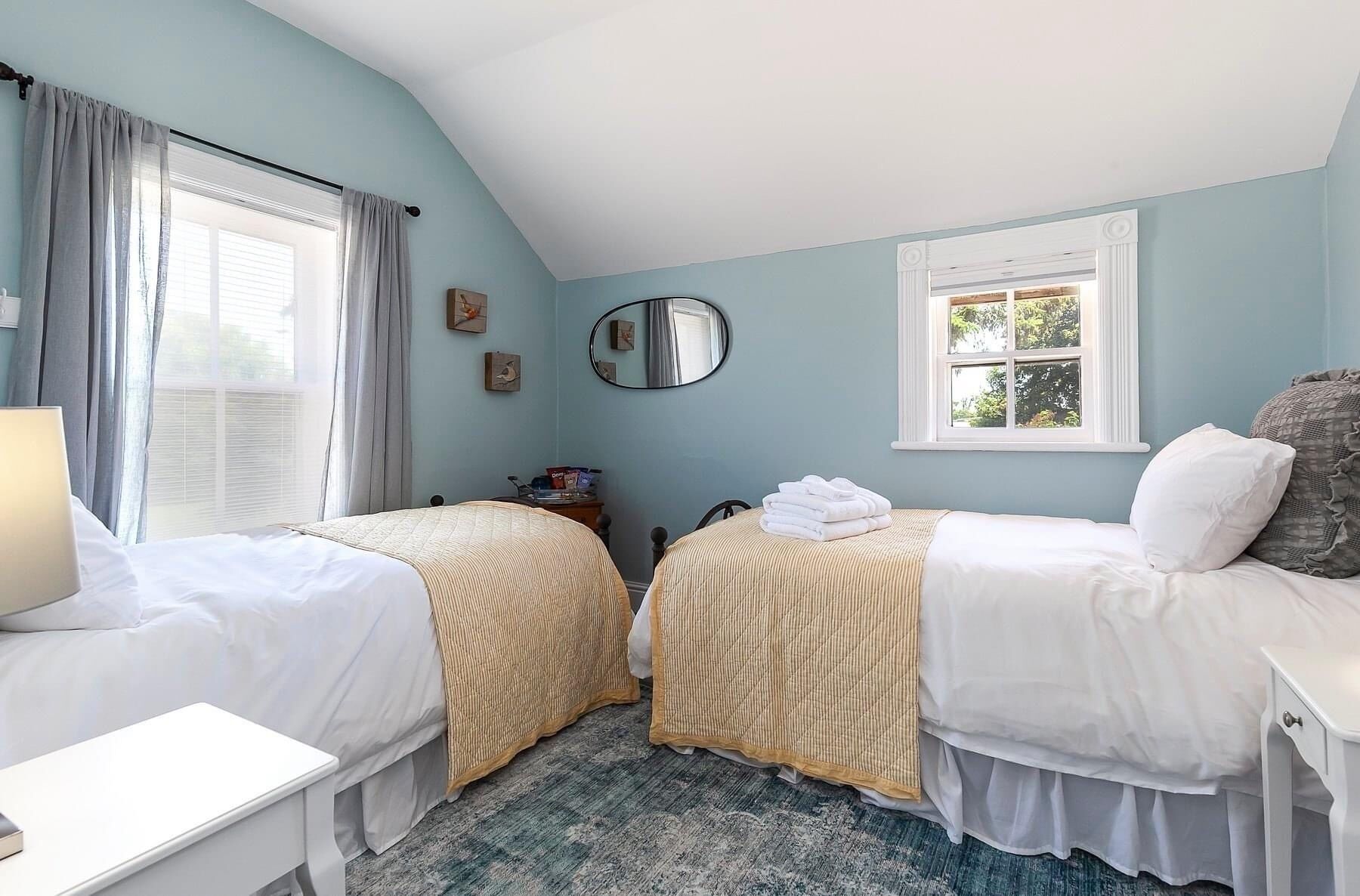 Een slaapkamer met twee bedden, een nachtkastje, een lamp, een raam met gordijnen en een spiegel aan de muur.