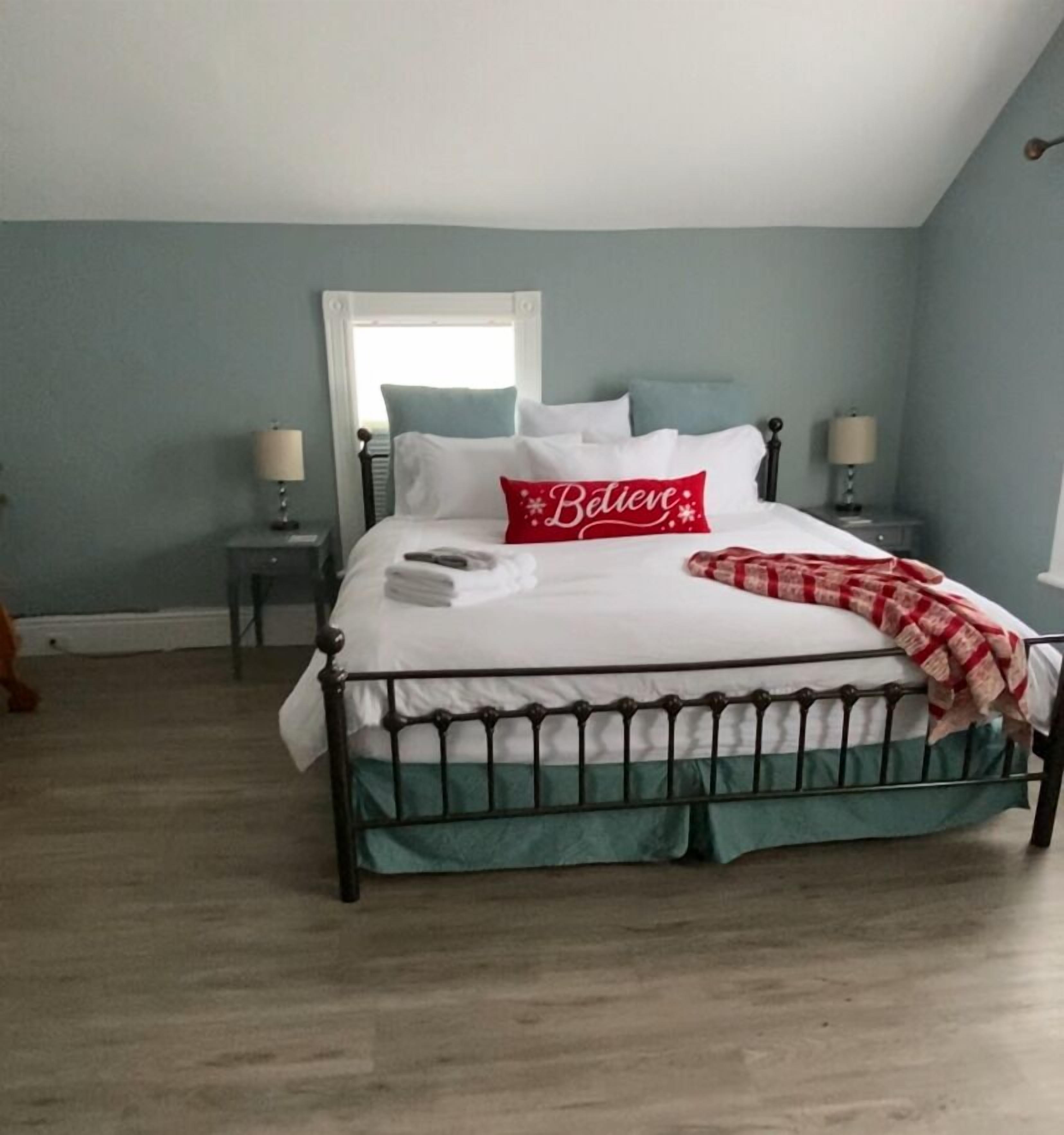 Una cama bien hecha con sábanas blancas, un cojín rojo con la palabra “Believe” y una manta a cuadros. La habitación tiene paredes azul claro, una ventana con cortinas blancas y un suelo de madera.