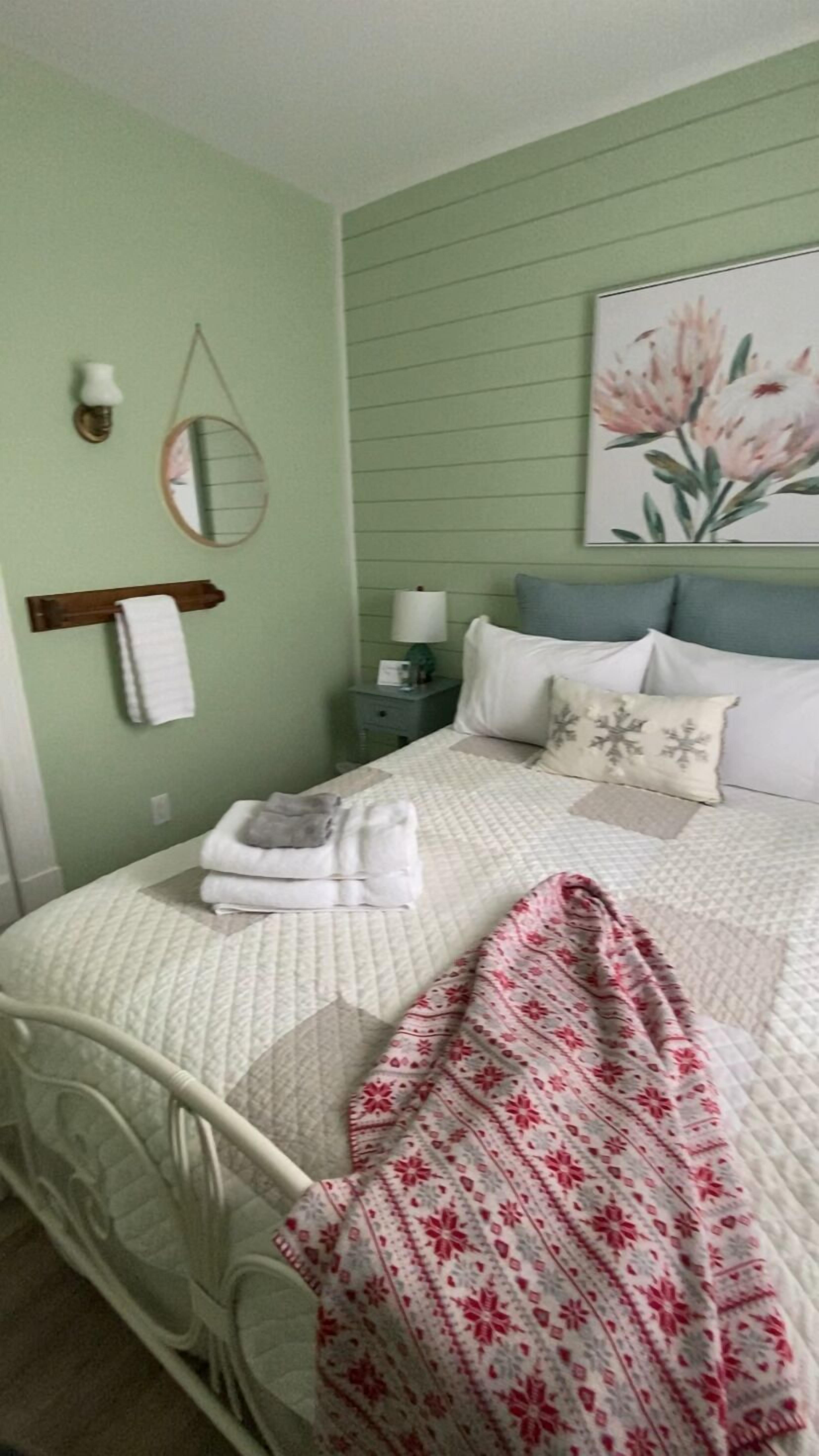 Una cama bien hecha con almohadas y una colcha, una mesita de noche con una lámpara, un espejo en la pared y un cuadro de flores.