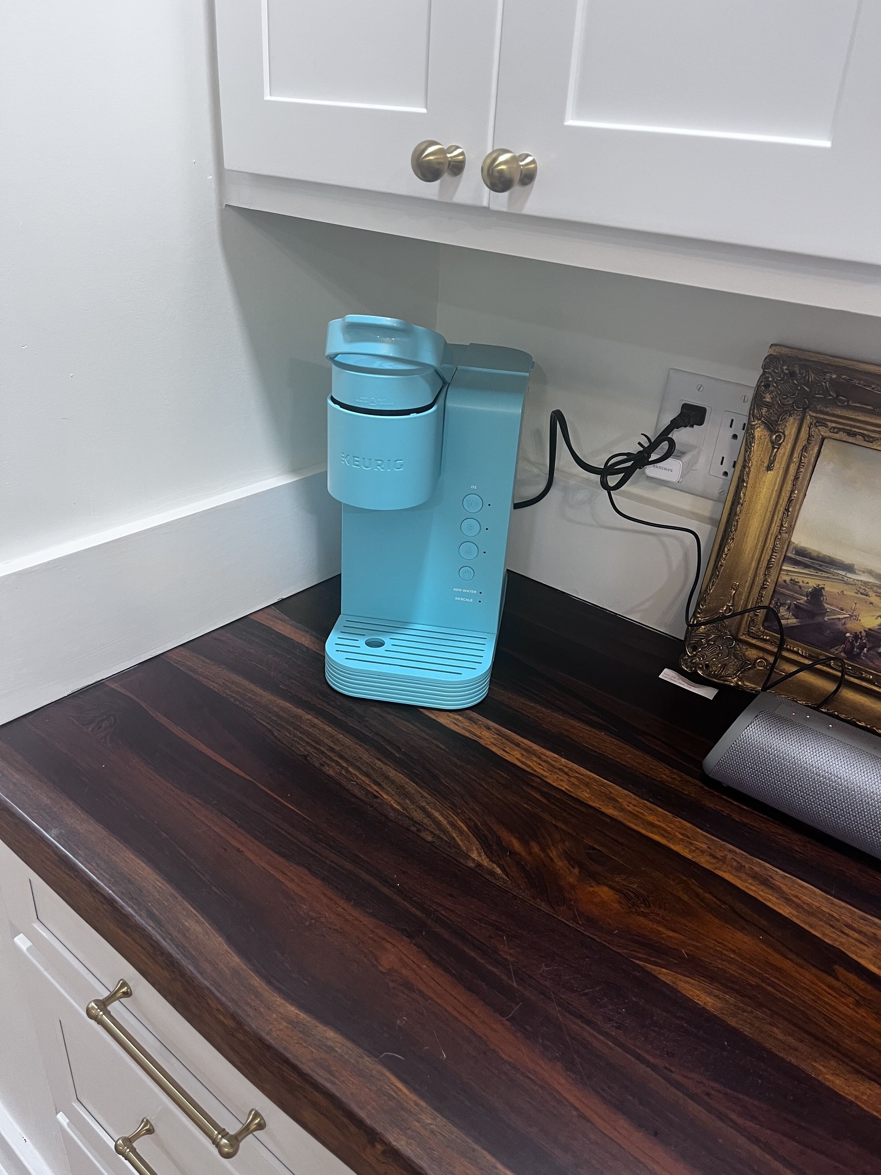 A teal Keurig coffee maker on a dark wooden countertop, connected to a power outlet.