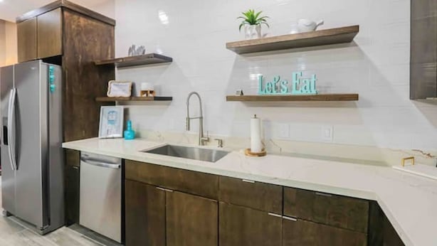 Eine moderne Küche mit dunklen Holfächern, einem Edelstahl-Kühlschrank, einer Spülmaschine und einer weißen Arbeitsplatte. Oberhalb der Arbeitsplatte befinden sich zwei Holzböden, einer mit einer Pflanze und der andere mit einem dekorativen Gegenstand.