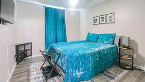 Ein Schlafzimmer mit Bett, kleinem Tisch, Stuhl, Fenster mit Vorhängen und einer Wandablage.