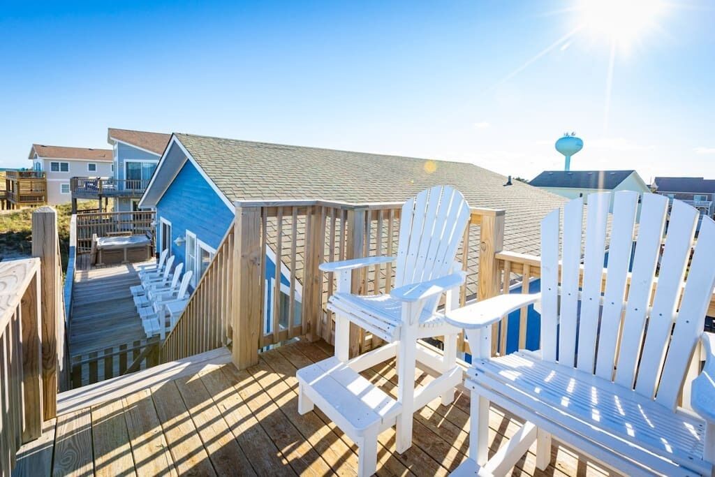 A wooden deck with white Adirondack chairs, a blue building, and a clear sky.