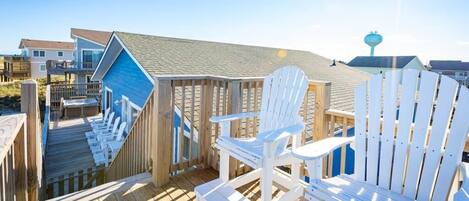 A wooden deck with white Adirondack chairs, a blue building, and a clear sky.