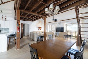 A spacious dining area with a wooden table and chairs, a ceiling fan, and a view of the living room with a sofa and television. - New 2026 Renovations Retreat Just Steps to Beach! (Kitty Hawk)
