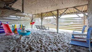 A covered beach area with a sandy floor, a children's play area with slides and a small boat, and a swing set. - New 2026 Renovations Retreat Just Steps to Beach! (Kitty Hawk)