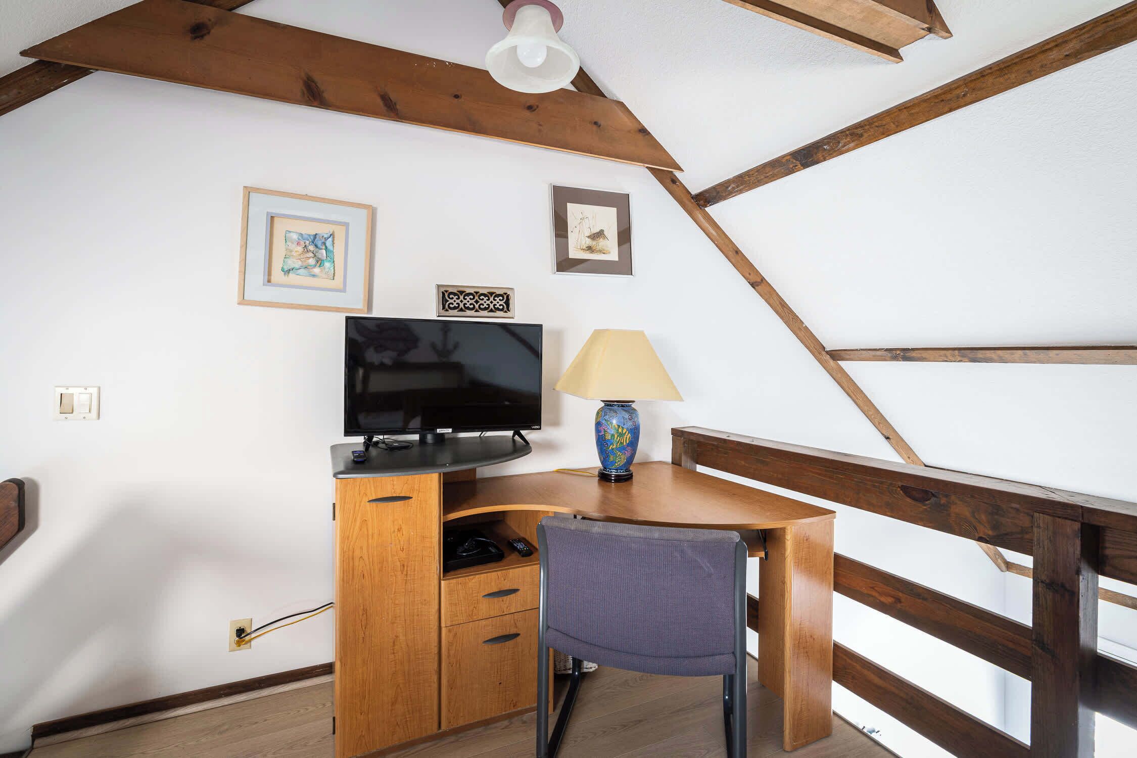 A wooden desk with a television, a lamp, and a chair in a room with exposed wooden beams and white walls.