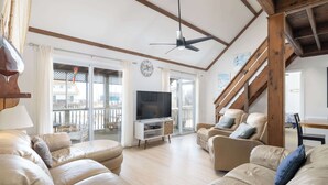 A spacious living room with a large TV, comfortable leather sofas, a wooden ceiling fan, and a balcony with a view. - New 2026 Renovations Retreat Just Steps to Beach! (Kitty Hawk)