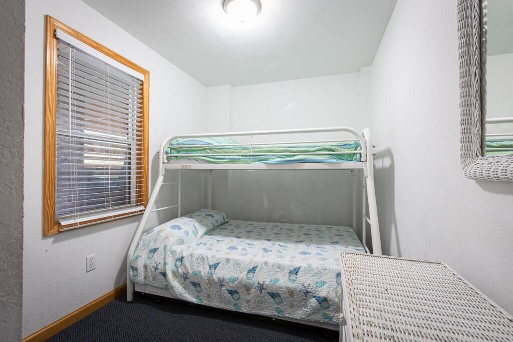 A small, single-bed room with a bunk bed, a window with blinds, a woven chair, and a mirror on the wall.