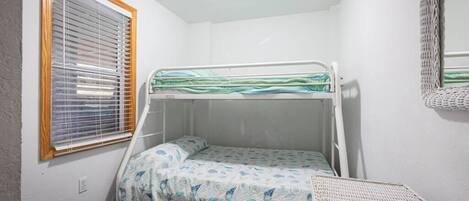 A small, single-bed room with a bunk bed, a window with blinds, a woven chair, and a mirror on the wall.