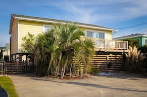 A single-story yellow house with a wooden deck, surrounded by palm trees and a wooden fence. - Near Beach, Dining & Shops, Private Hot Tub (Kill Devil Hills)
