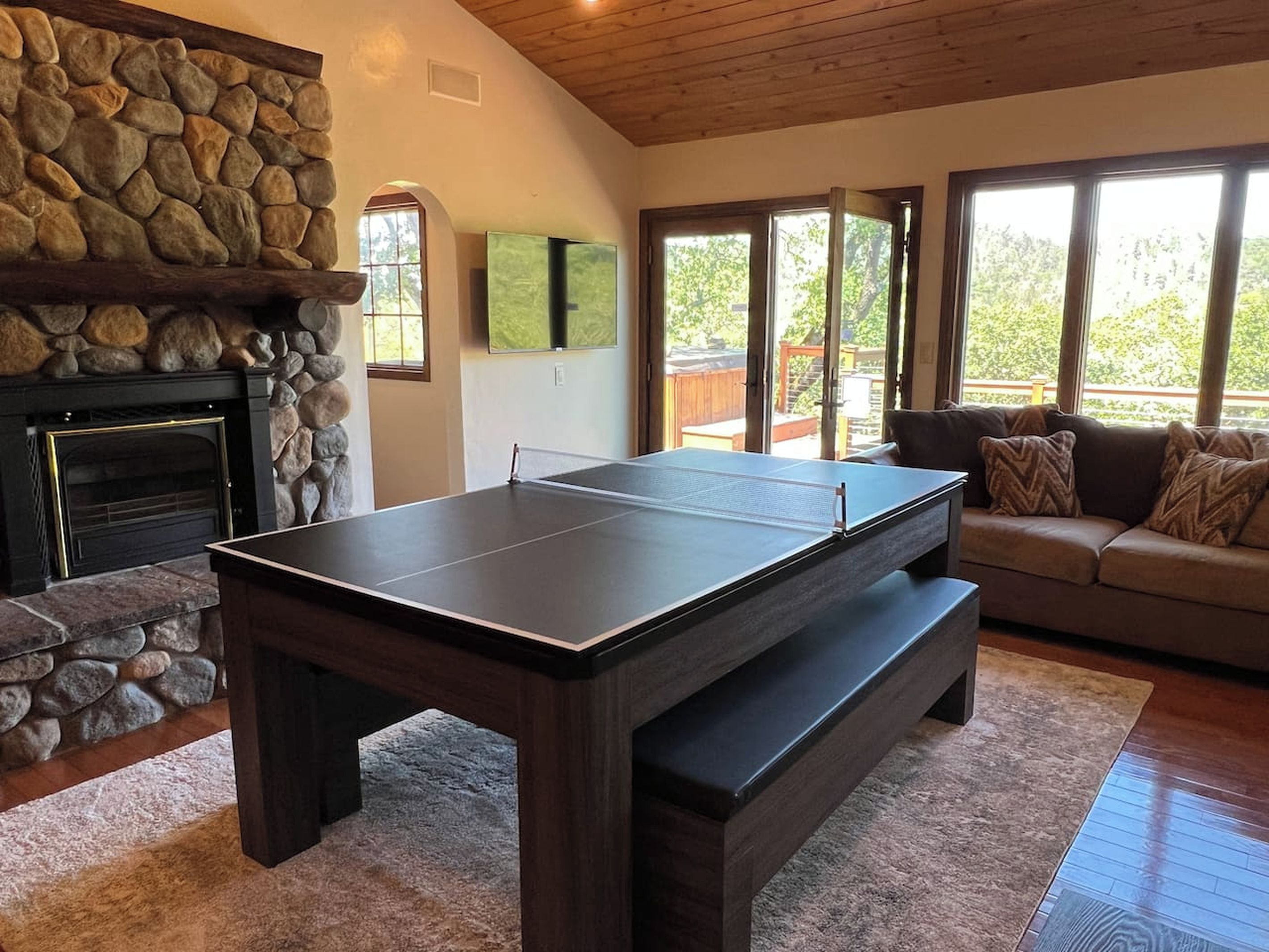 Une table de ping-pong dans une pièce dotée d’une cheminée en pierre, d’un canapé et de grandes fenêtres.