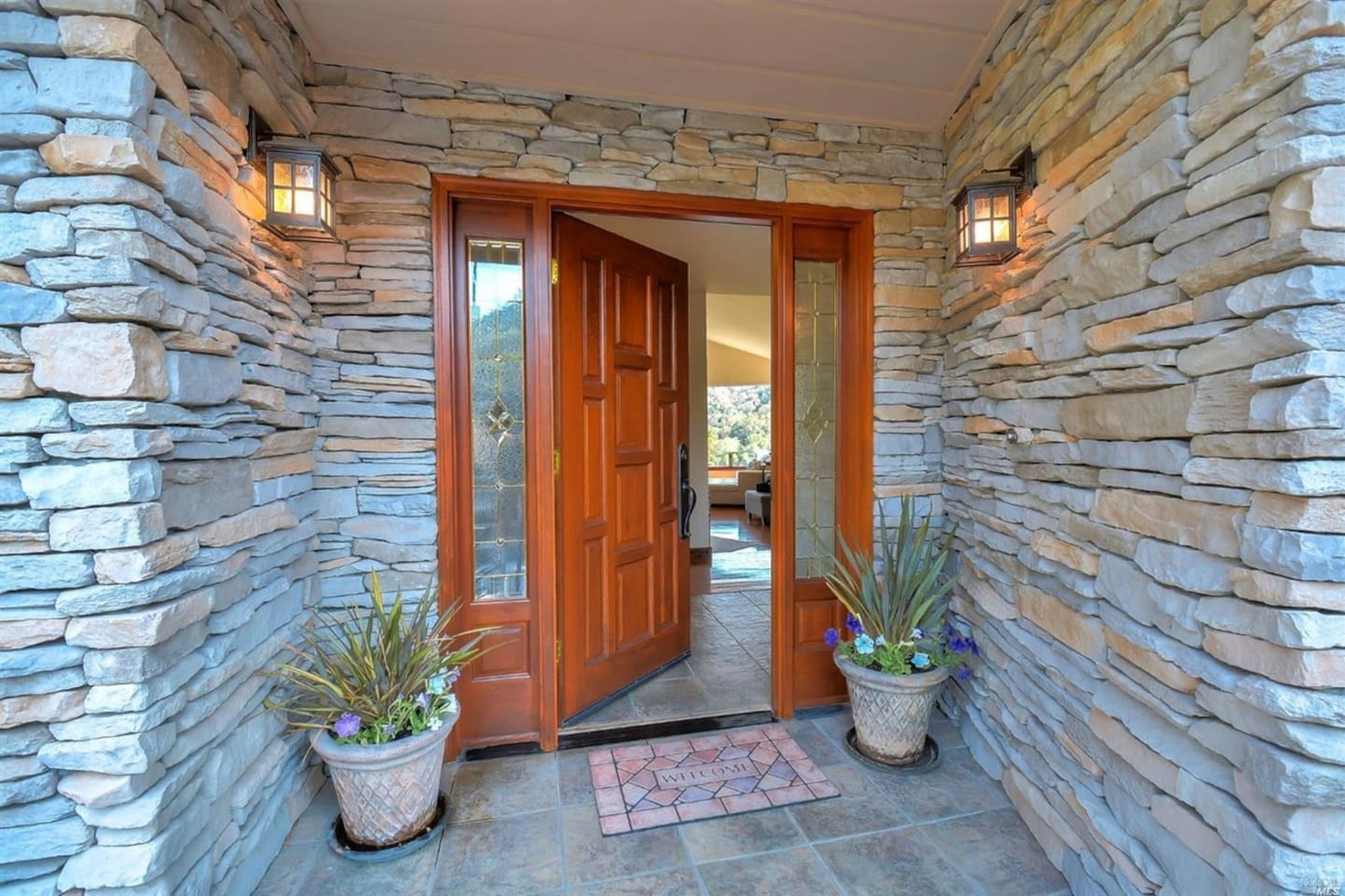 A stone-walled entrance with a wooden door, potted plants, and a welcome mat.