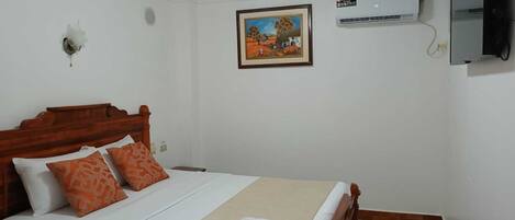 Una cama bien hecha con una colcha color beige y dos almohadas naranjas, un cuadro en la pared y un aire acondicionado de pared.