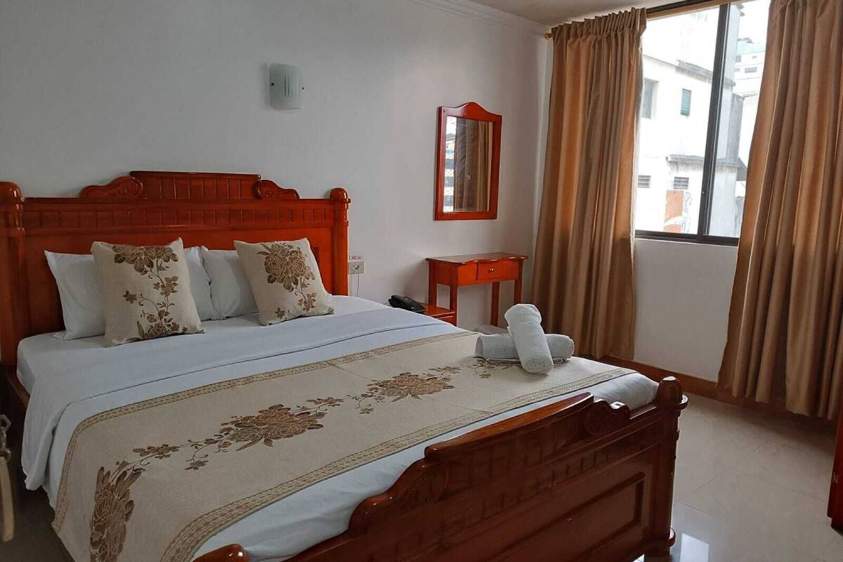 A hotel room with a wooden bed, a small bedside table, a mirror, and a window with curtains.