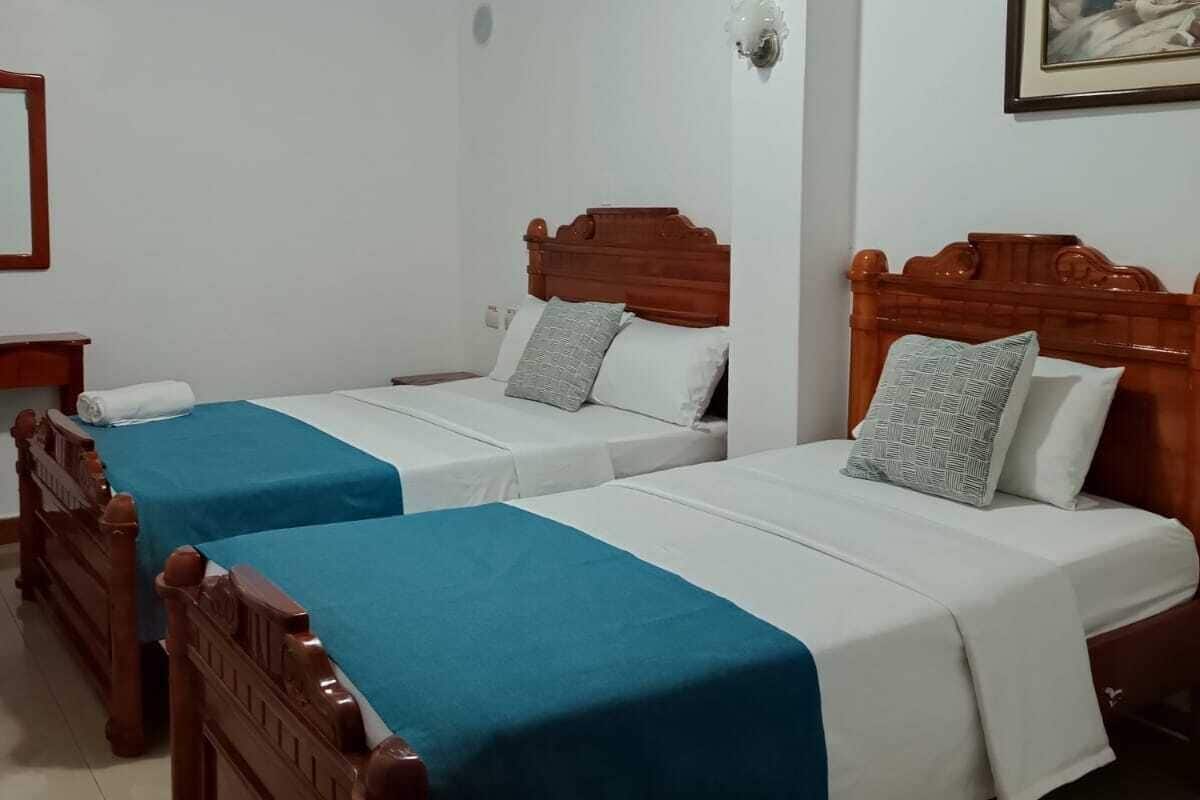 A hotel room with two wooden beds, each with a blue bedspread and white linens. There is a ceiling light fixture and a wall-mounted light.