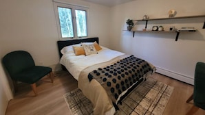 Un dormitorio con una cama, dos sillas, un ventanal y una estantería con objetos decorativos.