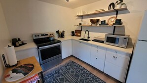 Una cocina compacta con armarios blancos, un microondas, una estufa y un lavabo. Hay repisas sobre la encimera que sostienen distintos utensilios de cocina.